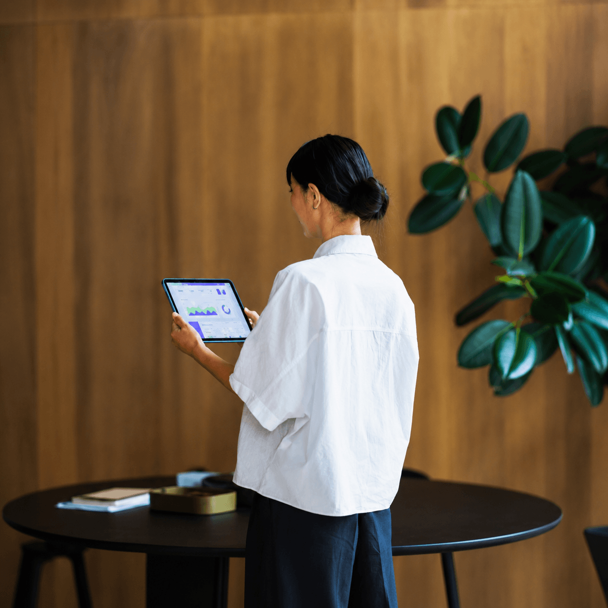 Woman viewing an analytics dashboard on a tablet, seen from behind against a warm wood-panelled wall